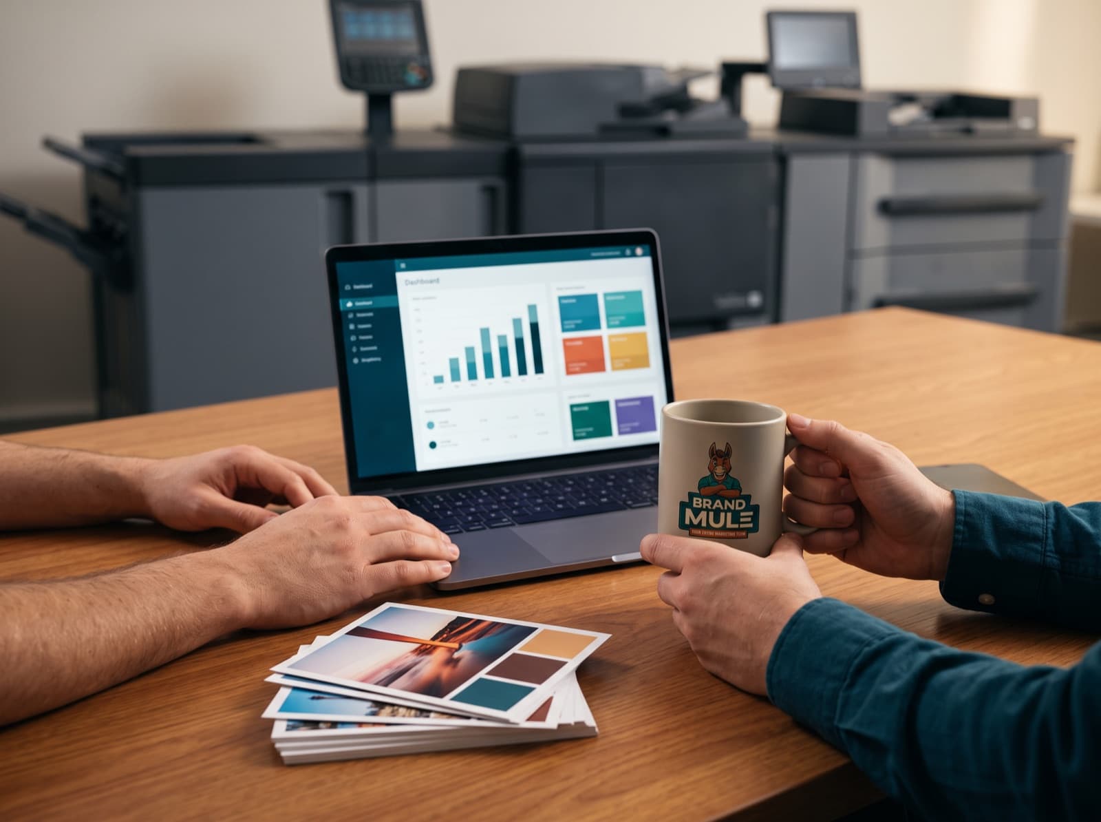 Two people at a table with a laptop showing a bar-chart dashboard, a stack of printed postcards and a Brand Mule mug between them, presses behind
