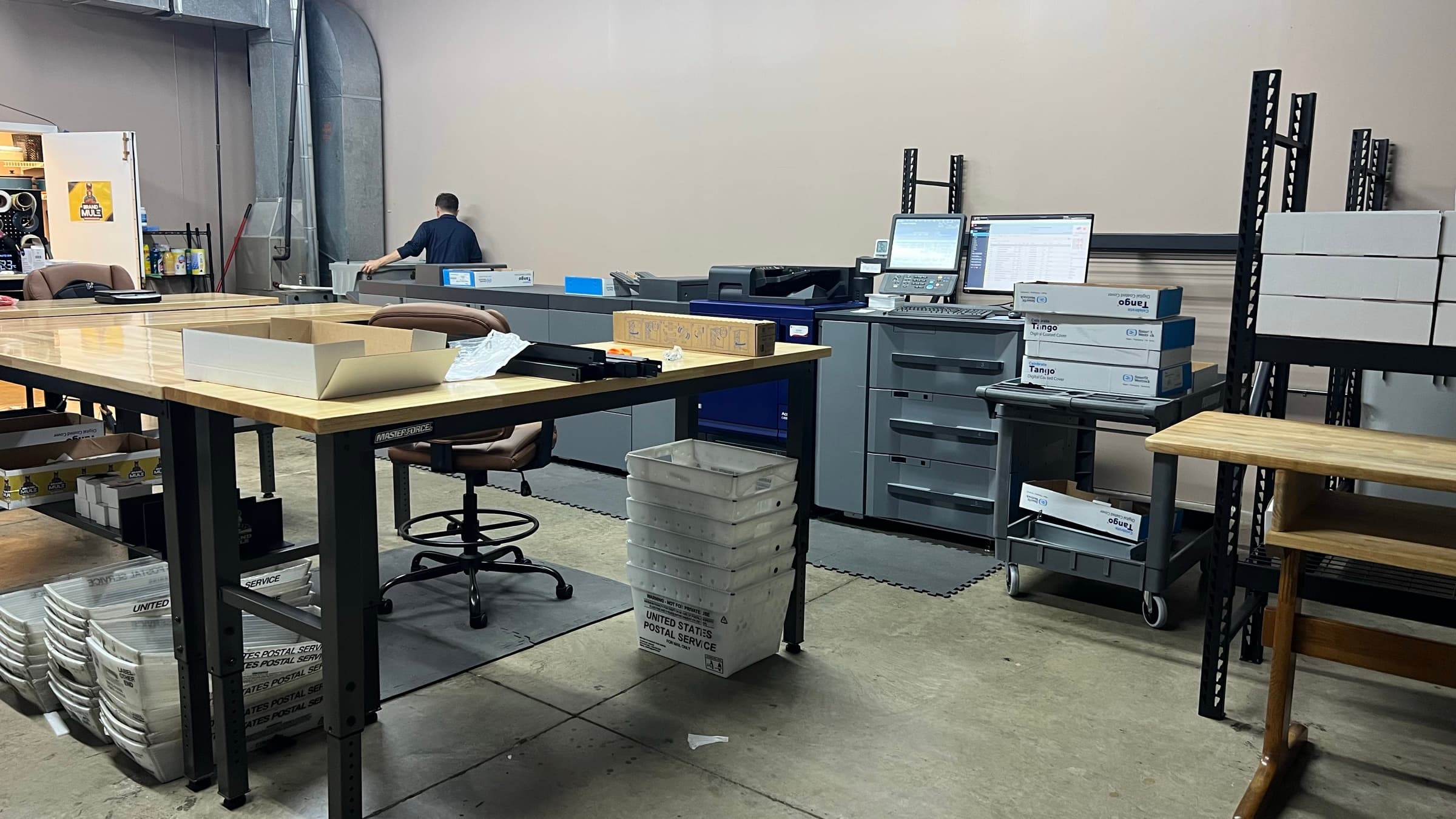 The shop floor: work tables, USPS mail trays and the press line with a team member at the controls