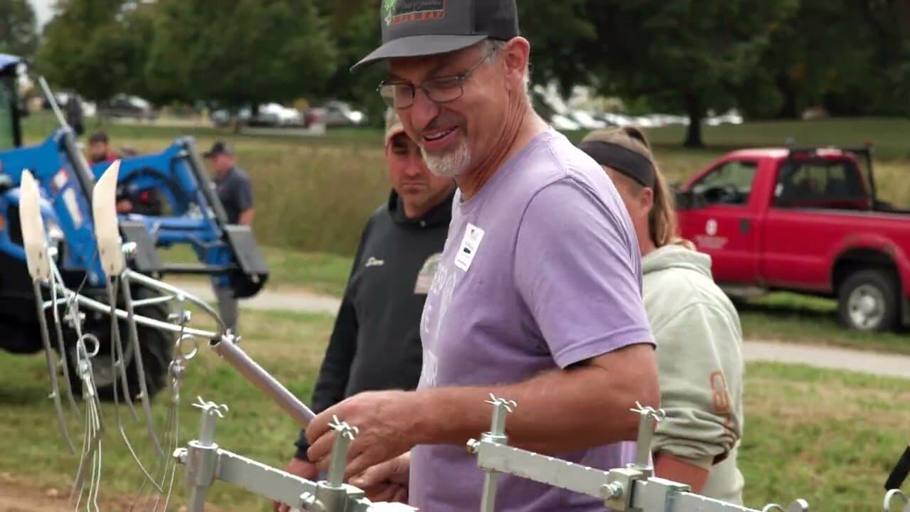 A smiling farmer at a tractor-mounted cultivator, a crowd and pickup trucks on the field behind