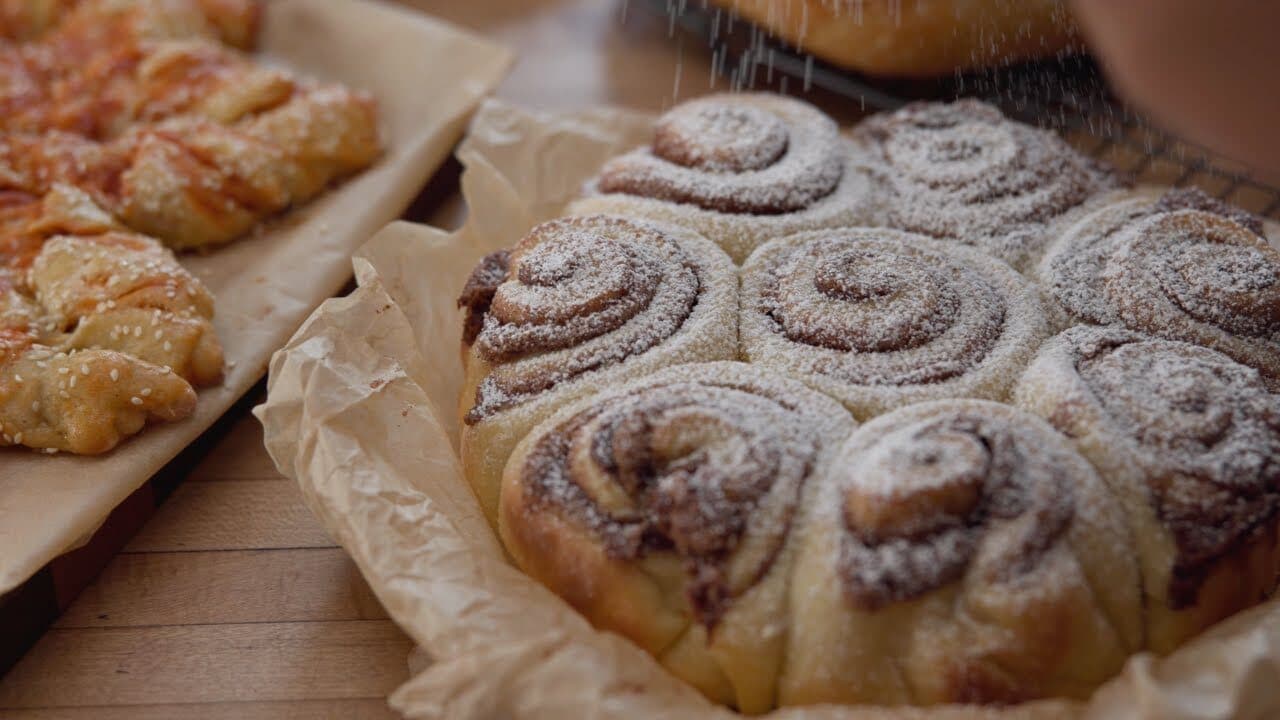 A tray of cinnamon rolls being dusted with powdered sugar, pastries on parchment beside them